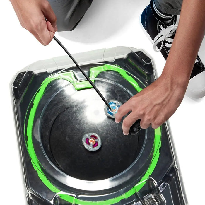 A person is launching a blue Beyblade from the Hasbro Beyblade X Xtreme Battle Set with Beystadium and 2 Right-Spinning Top Toys into the round, black arena using a ripcord. Another red spinning top from the set is already spinning inside the arena. The edge of the Beystadium features a green track. The person is wearing grey pants and black sneakers with white laces.