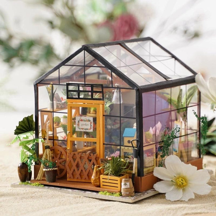 A detailed Rolife Miniature Cathy's Flower House with a transparent glass roof and walls, black framing, and a vibrant orange door. Inside, it is filled with tiny potted plants and gardening tools. The exterior is adorned with additional potted plants and flowers. This dream garden house rests on a sandy surface with white flowers around.