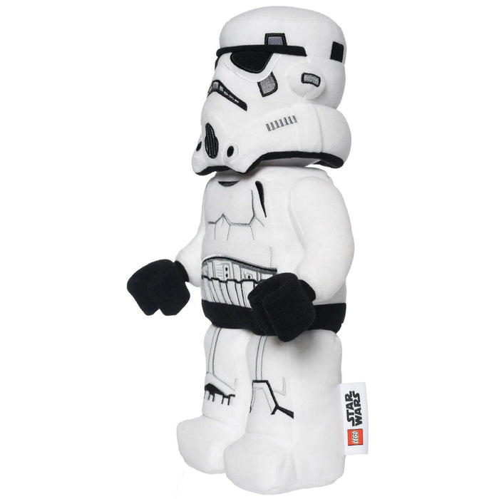 A plush toy shaped like a LEGO Star Wars Stormtrooper is shown. The LEGO Star Wars: Stormtrooper Plush Minifigure by Little Shop of Magic is predominantly white with black details outlining the helmet, body armor, and gloves. It stands upright, and a small tag with LEGO and Star Wars logos is attached to its leg.