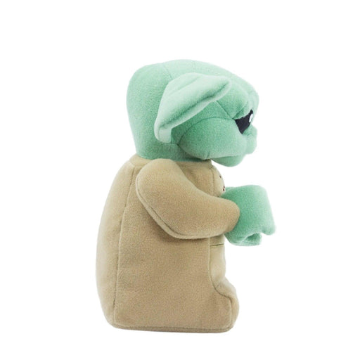 A side view of a plush toy resembling a green, large-eared, alien-like character from The Mandalorian with a beige robe. The LEGO Star Wars: The Child Plush Minifigure by Little Shop of Magic is standing upright and has small, black eyes. It has a rounded head with prominent ears sticking out, and the beige robe covers its body, leaving only the green hands visible.