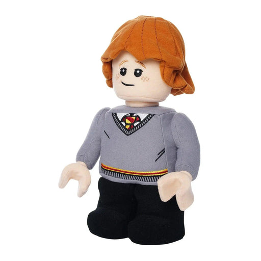 A Little Shop of Magic LEGO Harry Potter: Ron Weasley Plush Minifigure resembling a minifigure character with shaggy red-orange hair and a freckled face. The figure is dressed in a Gryffindor gray school uniform sweater with an embroidered emblem, a white collar, and a striped tie, paired with black pants. The toy has a smiling expression.