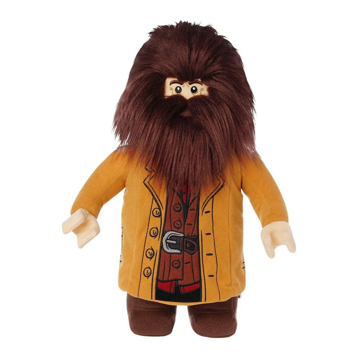 A plush character resembling the LEGO Harry Potter: Hagrid Plush Minifigure from Little Shop of Magic features a tall, bushy dark brown beard and long hair. The figure is dressed in a tan coat over a red vest and brown pants, with black buttons and a belt printed on the outfit. The toy has light-colored hands and a friendly, cartoonish facial expression.