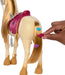 A hand places a colorful flower-shaped clip onto the long, blonde tail of an interactive toy horse. The Barbie Mysteries: The Great Horse Chase Interactive Toy Horse by Mattel has a light beige body, a pink saddle, and grey stirrups. The tail is already adorned with similar pink, blue, and purple clips as the horse faces away from the camera.