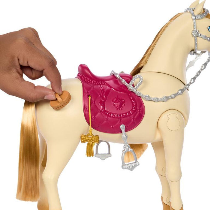 A hand is adjusting a tan knob on an interactive toy horse's side. The cream-colored Barbie Mysteries: The Great Horse Chase Interactive Toy Horse by Mattel boasts a shiny, golden mane and tail. It wears a pink saddle adorned with decorative details, including a star emblem, secured with a silver bridle. Two silver stirrups and a golden ribbon hang from the saddle.