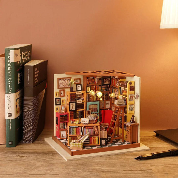A meticulously crafted Rolife DIY Miniature Dollhouse- Sam's Study of a cozy, cluttered library scene. It features a red armchair, numerous bookshelves filled with books and miniature decorations, a ladder, and warm lighting. The Rolife DIY Miniature Dollhouse- Sam's Study is placed on a wooden desk beside a lamp, tall books, and a notebook.
