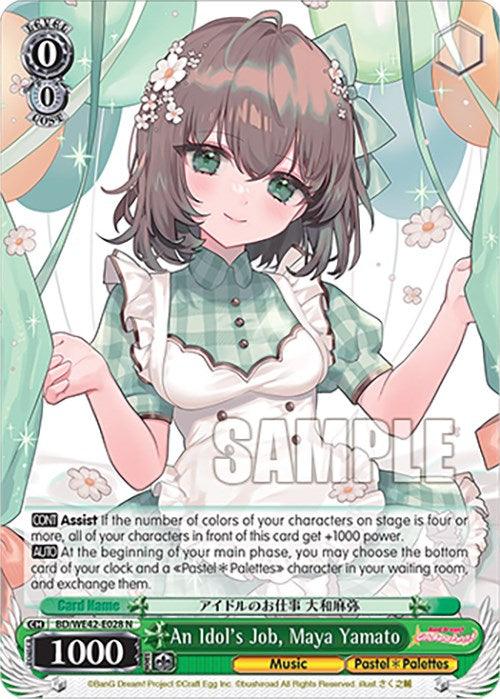 A trading card from the Bushiroad collection featuring an anime-style character, a young woman with short brown hair. She wears a green and white checkered dress with puffed sleeves and a white collar, holding up two fingers in a peace sign. The background is adorned with ribbons and flowers, while text overlays provide game stats and abilities. This card is titled "An Idol's Job, Maya Yamato (BD/WE42-E028 N) [BanG Dream! Girls Band Party! Countdown Collection].
