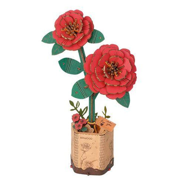 The Rowood Red Camellia by Robotime is a wooden handcrafted model that showcases two large, detailed red flowers with multiple layers of petals. Accompanied by green leaves, the flowers are displayed in a decorated pot adorned with engraved floral designs and a small tag that reads "FOR YOU." The overall design is intricate and artistic.