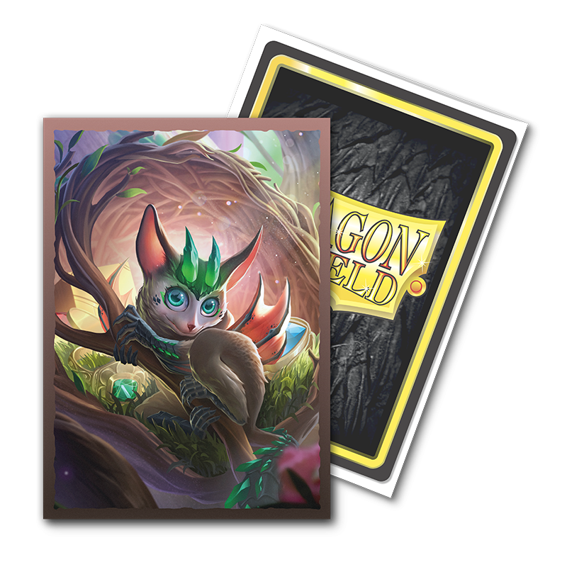 A fantasy trading card showcases a whimsical creature with large blue eyes, pointed ears, and a green leafy headdress, set against the backdrop of a vibrant forest with soft lighting. The textured back proudly displays "Dragon Shield" in yellow, as part of the Arcane Tinmen's TCG cards series. It's perfect for fans of Brushed Art sleeves and appropriately named "The Bushdrake.