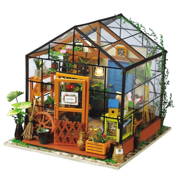 A Rolife Miniature Cathy's Flower House featuring a mix of plants, flowers, and gardening tools. This dream garden house by Rolife has transparent glass walls and a black frame. Inside, there's a blue door, wooden shelves with various potted plants. The entrance boasts a small orange gate, a "garden" sign, and a birdhouse.