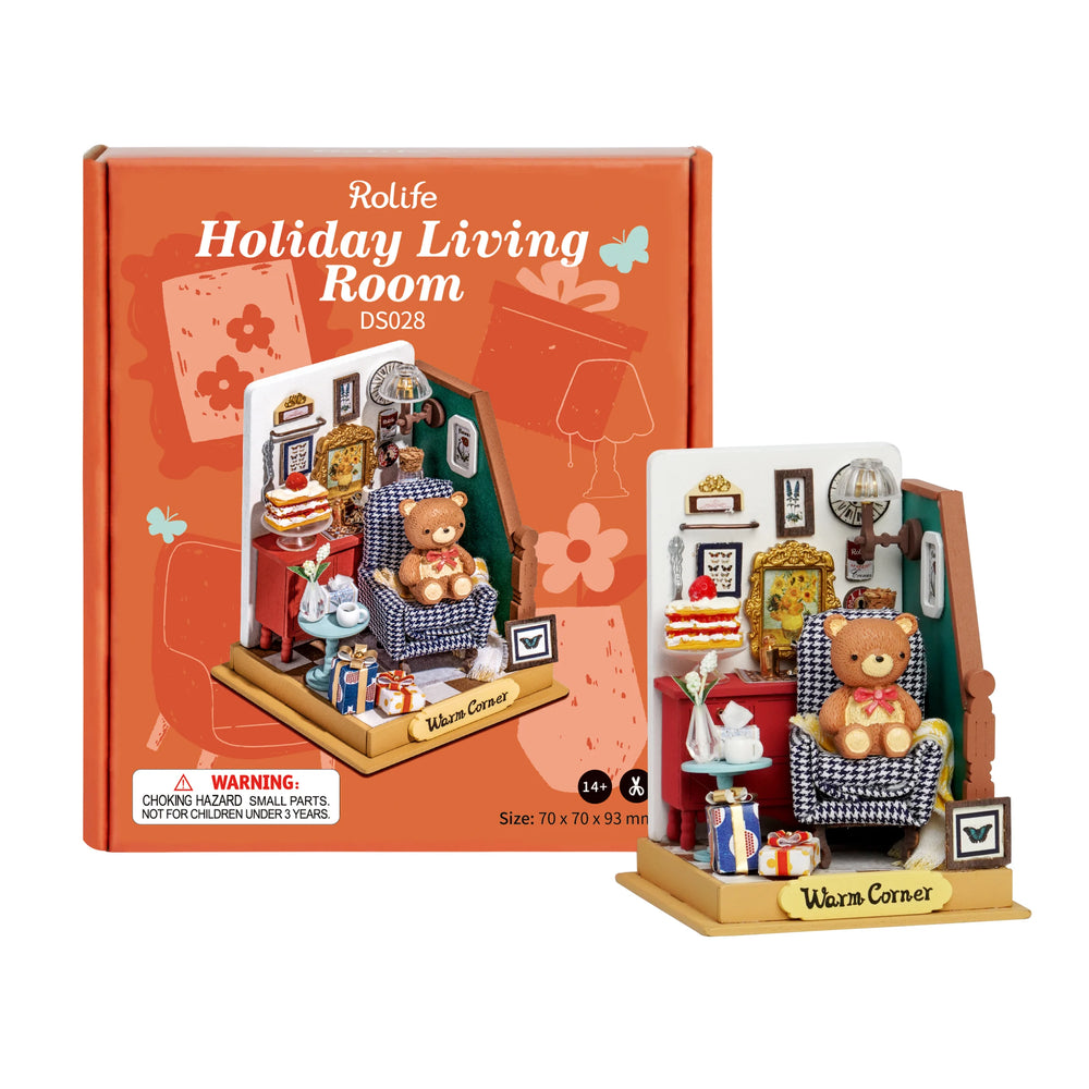 A miniature diorama kit titled "Holiday Living Room" by Robotime is displayed against an orange box. The scene, perfect for a relaxing weekend, includes a teddy bear seated in a checkered armchair within a cozy home atmosphere featuring framed art, a Christmas tree, gifts, and small furniture. The kit is marked for ages 14+ and measures 70 x 70 x 93 mm.