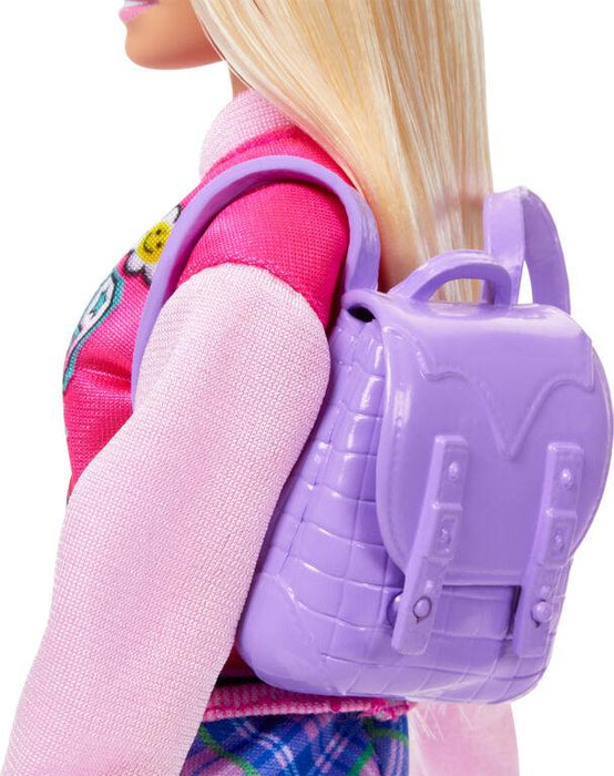 A close-up view of a doll with long blonde hair wearing a pink shirt with a decorative patch on the right sleeve and a purple backpack. The backpack, part of the Barbie-I Love School-Blonde Doll by Mattel accessories, has a quilted texture and buckle details. The doll's head is turned slightly to the left, and only part of the face is visible.