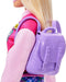A close-up view of a doll with long blonde hair wearing a pink shirt with a decorative patch on the right sleeve and a purple backpack. The backpack, part of the Barbie-I Love School-Blonde Doll by Mattel accessories, has a quilted texture and buckle details. The doll's head is turned slightly to the left, and only part of the face is visible.
