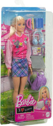 A Mattel Barbie-I Love School-Blonde Doll in a pink jacket adorned with various patches, a blue patterned skirt, and white shoes is packaged alongside back-to-school accessories. Included in the playset are a laptop, headphones, a water bottle, a pink school bag, and a purple backpack, all against a school-themed backdrop.