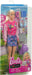 A Mattel Barbie-I Love School-Blonde Doll in a pink jacket adorned with various patches, a blue patterned skirt, and white shoes is packaged alongside back-to-school accessories. Included in the playset are a laptop, headphones, a water bottle, a pink school bag, and a purple backpack, all against a school-themed backdrop.