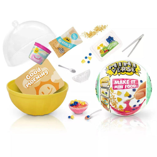 A yellow plastic bowl filled with a small egg-themed "Good Morning" card is surrounded by tiny food items including granola, colorful fruit, a spoon, tiny tweezers, a packet, a transparent dome lid, yogurt, and a wrapped MiniVerse ball labeled "MGA Make It Mini Food Cafe Series 3 Mini Collectibles.