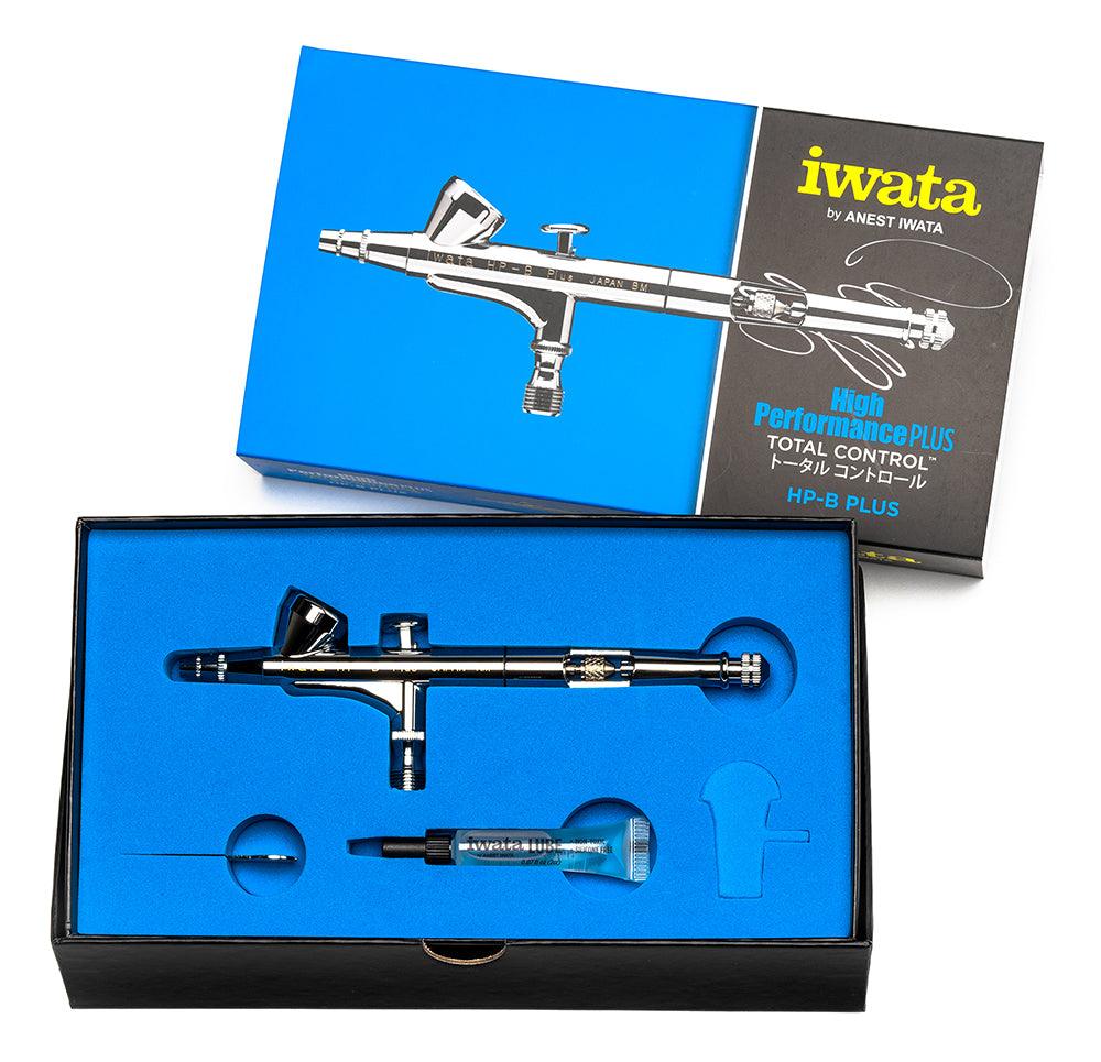 The Iwata High Performance HP-B Plus airbrush set showcases a sleek metal airbrush nestled in blue foam, with packaging featuring a black and silver illustration highlighting its precise control for detailed spraying. Accessories are securely stored within the foam compartments.