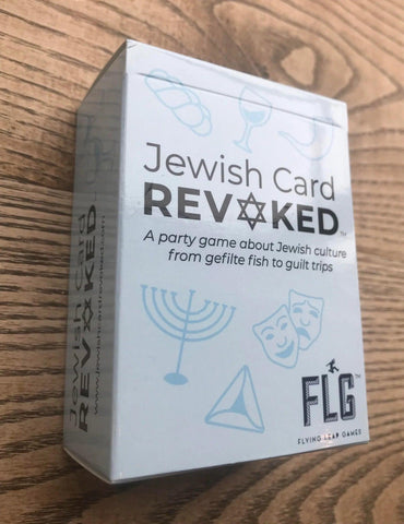 A rectangular box of the game "Jewish Card Revoked" is set against a wooden background. The box features text: "A party game about Jewish culture from gefilte fish to guilt trips." It is adorned with light blue icons including a wine glass, fish, comedy/tragedy masks, and a dreidel. The logo "FLG" and the brand name "Flying Leap Games" appear at the bottom.