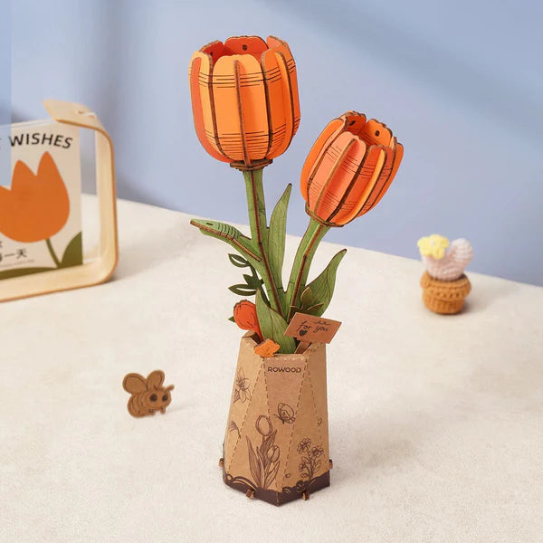 The Rowood DIY Wooden Flower Bouquet by Robotime features two orange tulips with green stems and leaves in a floral hexagonal cardboard vase, accompanied by a wooden mouse cutout and a cupcake figurine, with a card bearing an orange tulip illustration and 