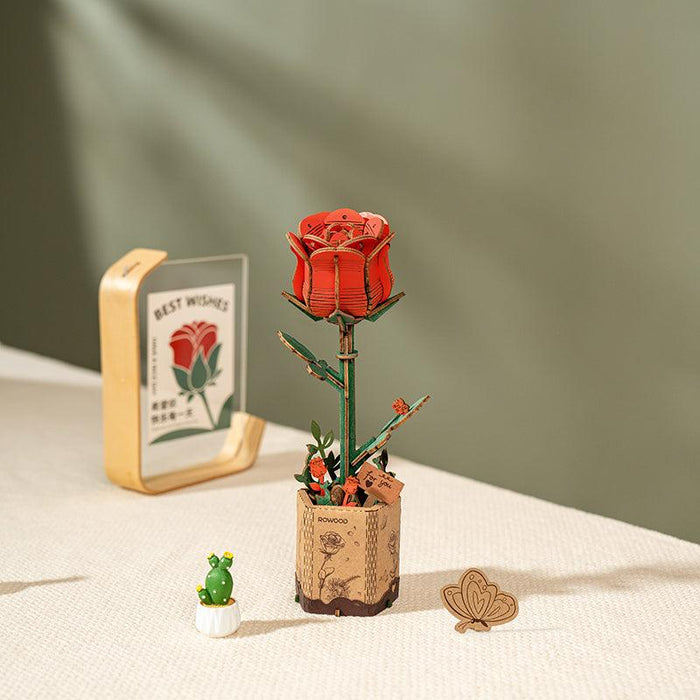 A decorative setup features a **Rolife Rowood Red Rose** in a vase on a beige surface. Nearby, there's a small white vase with a miniature green cactus, a wooden heart-shaped ornament boasting meaningful design, and a framed card with a rose illustration and the text "BEST WISHES." The background is gradient from light to dark green.
