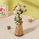 The Robotime Chamomile paper bouquet with white and yellow daisies, chamomile, and bees in a brown vase sits on the table. Nearby, a small brown bear figure and a tiny chick in a cup add charm. A book with red flowers is partially visible against the pale yellow wall.