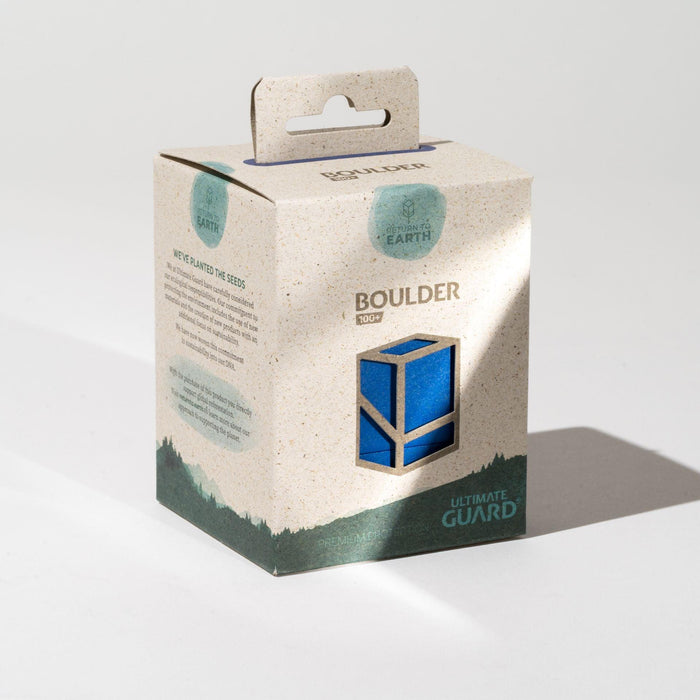 A beige cardboard box with green text, featuring the “RETURN TO EARTH BOULDER BLUE” sustainable deck case by Ultimate Guard. The box showcases a blue 3D cube design on the front and has an eco-friendly "Return to Earth" logo. A small cut-out window reveals part of the blue cube inside. A hanging tab is visible at the top.