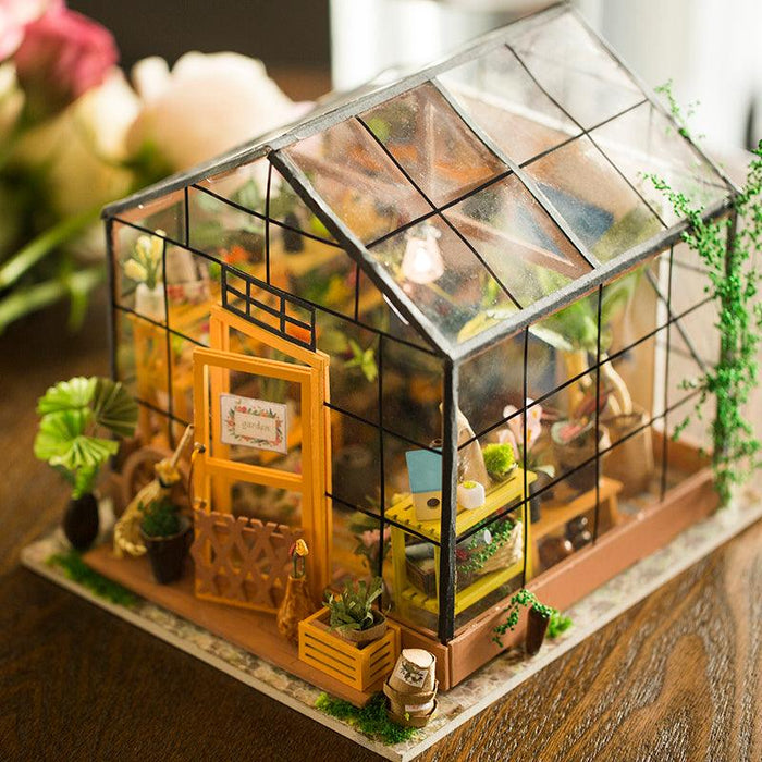 A dream garden house with a transparent glass structure and black frame sits on a wooden surface. The detailed interior includes tiny potted plants, a wooden bench with gardening tools, and a welcoming sign on the front door. Various greenery adorns the outside, creating a lively scene, as seen in the Rolife Miniature Cathy's Flower House by Rolife.
