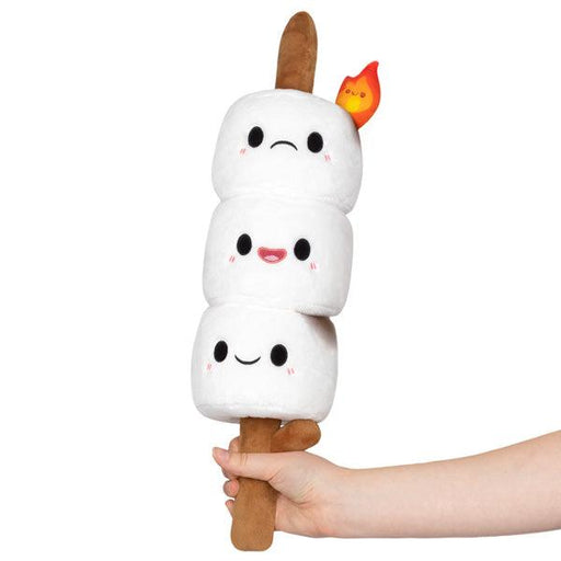 A hand holds a plush toy resembling a skewer with three mini marshmallows stacked on it. Each marshmallow has a face with black eyes and pink cheeks. The top one has a neutral expression, while the middle and bottom ones are smiling. A small, plush flame is attached near the top, perfect for camping-themed fun. This is the Squishable Mini Comfort Food Marshmallows.