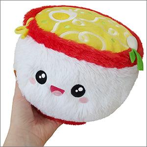 A hand holds the Mini Comfort Food Ramen Squishable® from Squishable, a plush toy designed to look like a fluffy bowl of pork tonkatsu ramen. With its red base, white furry middle, and yellow noodle-like top, this adorable product features a cute smiley face with big eyes and blush marks. It captures the charm of vibrant ramen restaurants in Shibuya Tokyo with playful green and yellow details.