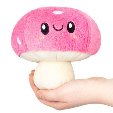 The Snugglemi Snackers Mushroom by Squishable is a plush toy with a pink cap and white stem. The cap has smiling black eyes, a happy mouth, and pink cheeks with yellow lines. Its soft fur stem fits perfectly in hand to showcase its round shape.