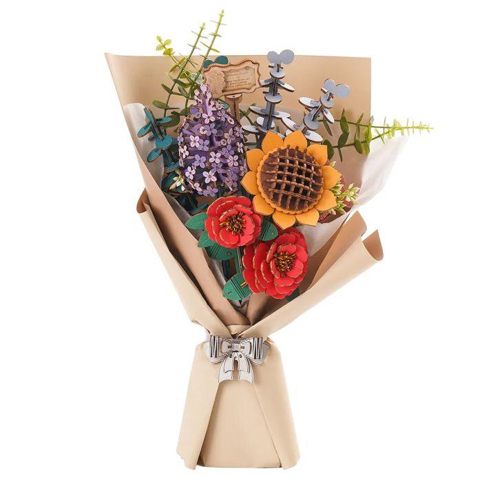 Introducing the Robotime Wooden Flower Bouquet, a stunning floral arrangement wrapped in elegant beige paper and tied with a silver ribbon. Featuring pre-colored blooms including sunflowers, red roses, purple flowers with green stems and leaves, and metallic gray ornamental accents. A small decorative plaque adds an extra touch of charm to this exquisite Robotime creation.