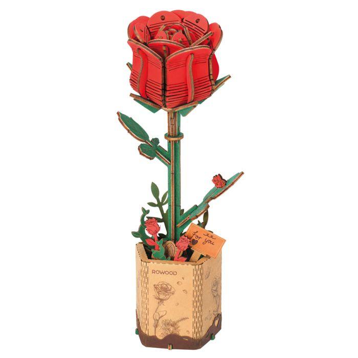 A 3D wooden puzzle model of a red rose atop a green stem with leaves stands in a beige, decorative base. The hexagonal base features illustrations of roses and includes a small tag reading 