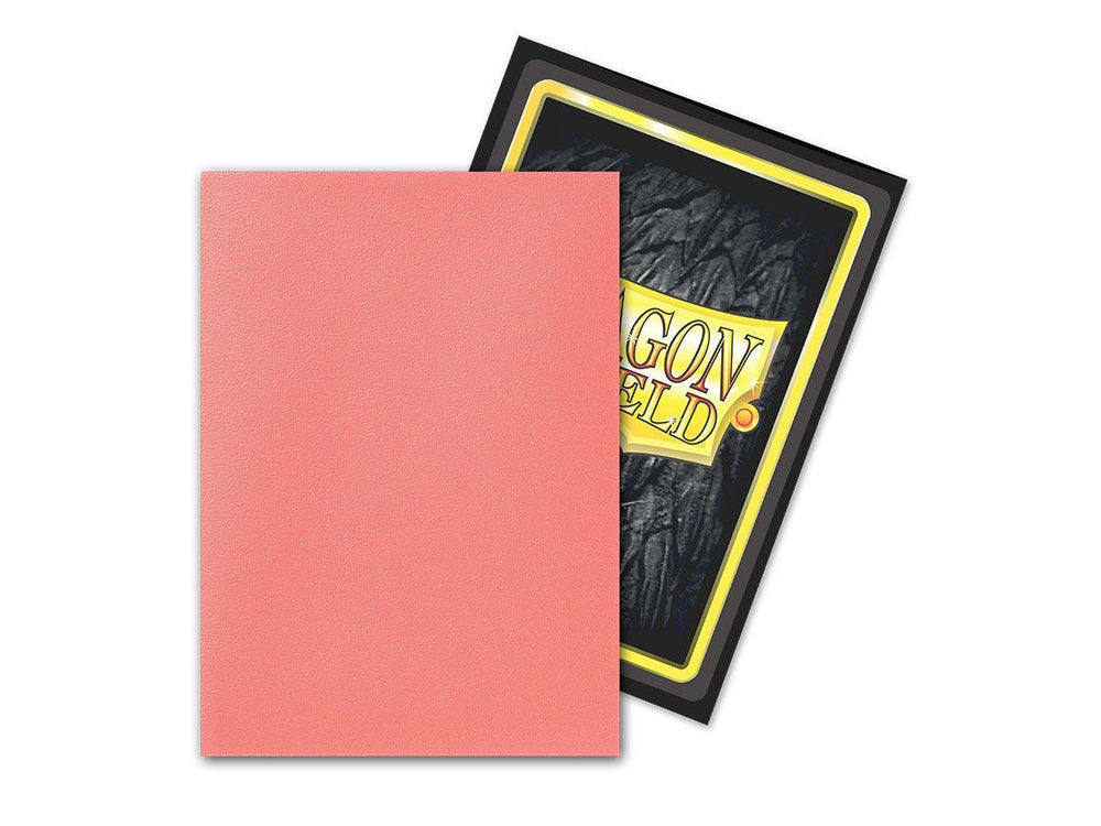 A single card sleeve is shown in the foreground, its backside facing the viewer and colored in a solid pink shade. Behind it, another card sleeve from the Arcane Tinmen Dragon Shield: Standard 100ct Sleeves - Peach (Dual Matte) collection is partially visible, featuring a design with black and yellow borders and the text "DRAGON SHIELD" on a rugged background.