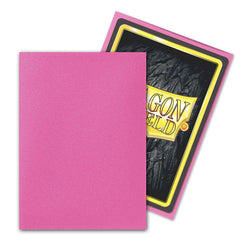 Two trading card sleeves are displayed. The pink one in the foreground, known as Dragon Shield: Standard 100ct Sleeves - Pink Diamond (Matte) by Arcane Tinmen, features a textured surface. In the background, a sleeve with a clear front reveals its back side with a black, stone-like design and a yellow text box that reads "DRAGON SHIELD.