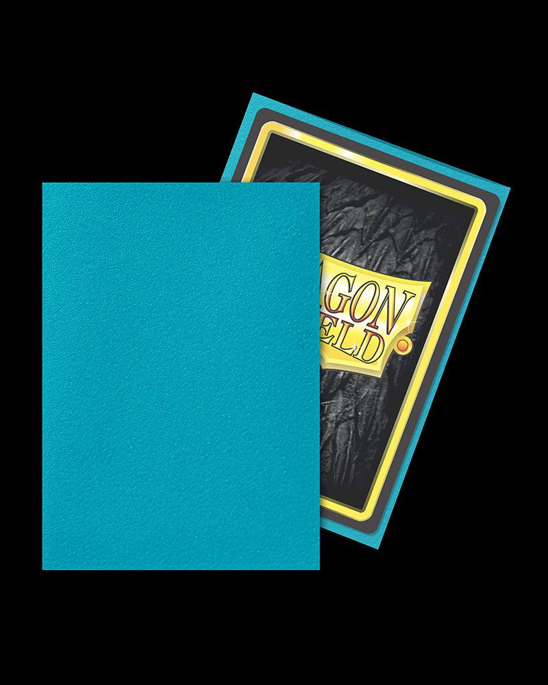 An image shows a card sleeve with a blue outer side and a partially visible card, which has a textured black background and a gold rectangular frame with the word "DRAGON" in large letters. The Arcane Tinmen Dragon Shield: Standard 100ct Sleeves - Turquoise (Matte) covers most of the card, leaving only the top part visible, perfect for TCG accessories enthusiasts.