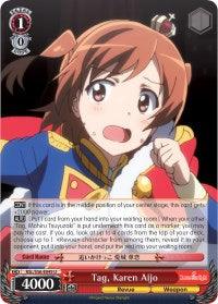 A trading card features an animated character from Revue Starlight, a young girl with short brown hair wearing a blue and red military-style outfit. She appears worried, holding a red paper in her left hand. The Tag, Karen Aijo (RSL/S56-E049 U) [Revue Starlight] by Bushiroad includes various stats and text, with "Tag, Karen Aijo" written at the bottom along with other game details.
