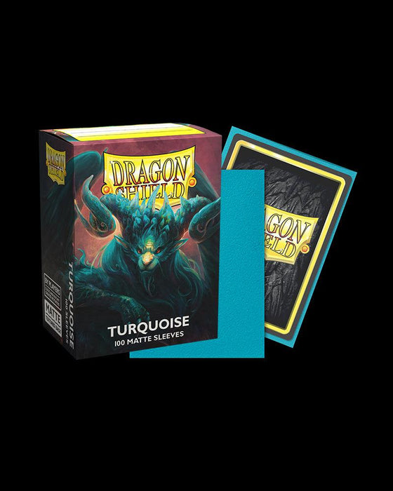 A box of Arcane Tinmen Dragon Shield: Standard 100ct Sleeves - Turquoise (Matte) trading card sleeves is displayed. The box features a dragon illustration. One turquoise sleeve is shown outside the box, and a card backed with a black and yellow Arcane Tinmen Dragon Shield card is partially inserted into another sleeve beside it, showcasing premium TCG accessories.