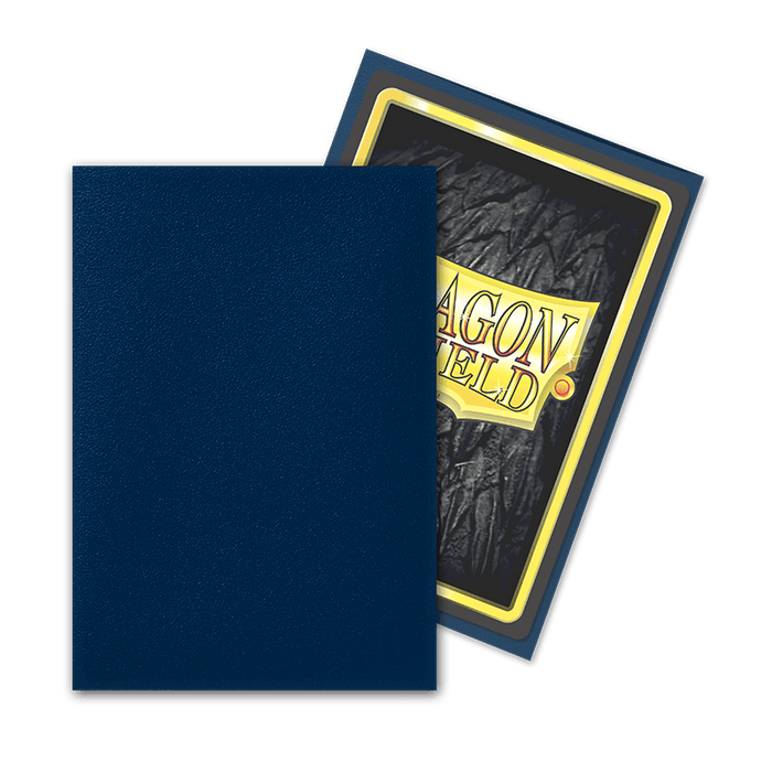 The image shows two Dragon Shield: Japanese Size 60ct Sleeves - Midnight Blue (Matte) by Arcane Tinmen. One sleeve is plain dark blue and is positioned in front of another sleeve that displays the back of a card featuring the "Dragon Shield" logo with a gold border, textured dark background, and yellow text. The card sleeves overlap slightly.
