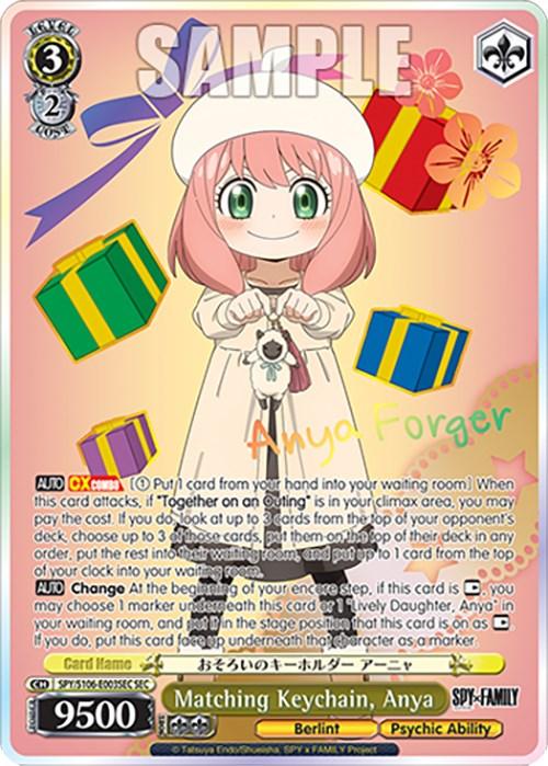 A Matching Keychain, Anya (SPY/S106-E003SEC SEC) [SPY x FAMILY] produced by Bushiroad. She has pink hair, green eyes, and is wearing a white dress with red shoes. Surrounded by colorful floating gift boxes, the keychain showcases her name and psychic abilities with various decorative elements and the series "SPY x FAMILY" indicated.