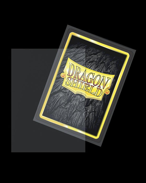 An Arcane Tinmen Dragon Shield: 100ct Outer Sleeves - Clear Matte (Standard) is shown with a black, textured background and a yellow border. The words "DRAGON SHIELD" are prominently displayed in golden letters on a banner design in the center. This Matte Clear Outer Sleeve offers excellent card protection, partially transparent and tilted slightly to the right.