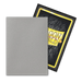 A trading card is displayed, with the front side turned away, showcasing a plain silver surface. Behind it, the back side of another trading card is partially visible, featuring the text "DRAGON SHIELD" against a dark, textured background with yellow and black borders—both protected by sleek Dragon Shield: Standard 100ct Art Sleeves - Justice (Dual Matte) from Arcane Tinmen.