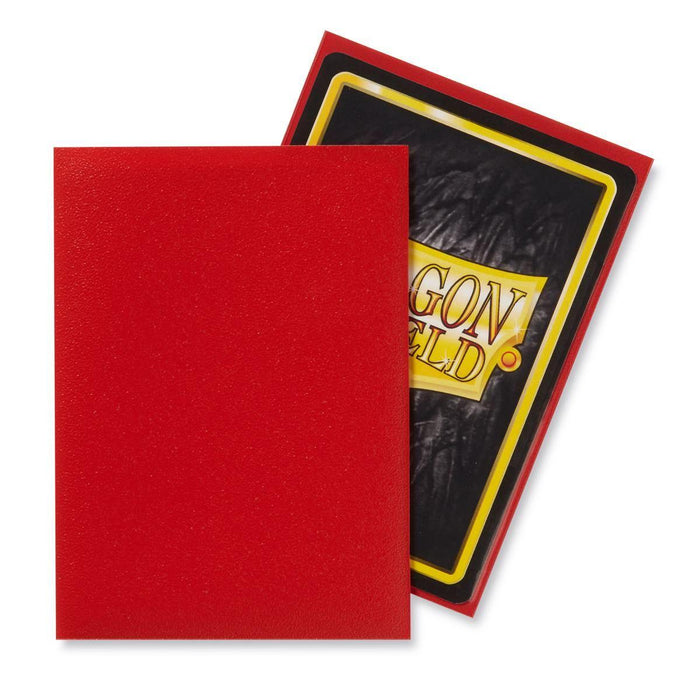 Two trading cards are shown. One card is face down, revealing its red back. The other card is partially scaled behind, displaying its matte sleeve with a black center and the text "DRAGON SHIELD" in yellow and red bordered in red from the Arcane Tinmen product Dragon Shield: Standard 100ct Sleeves - Crimson (Matte).