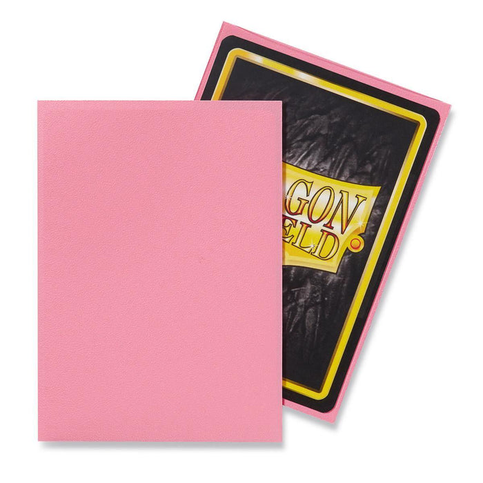 Two trading card sleeves are depicted. One sleeve shows the back of a trading card with a black background and partially visible yellow and red text that appears to say "DRAGON SHIELD." The other sleeve, which is solid pink, showcases Dragon Shield: Standard 100ct Sleeves - Pink (Matte). The items are arranged to display both the front and back of the sleeves from Arcane Tinmen.