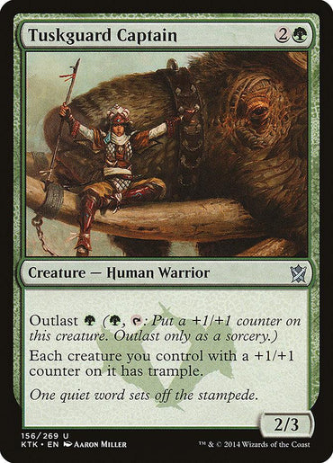 A Magic: The Gathering card named "Tuskguard Captain [Khans of Tarkir]," a Human Warrior from Khans of Tarkir, featuring artwork of a warrior standing next to a large beast. The card is green, costs 2G, and is a 2/3 creature with Outlast ability costing G and grants trample to other creatures. The flavor text reads: "One quiet word sets off the stamp.