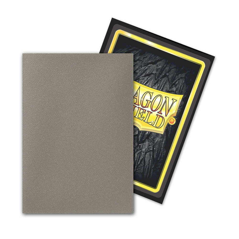 A textured, matte gray card is encased in Arcane Tinmen's Dragon Shield Japanese Size 60ct Sleeves - Crypt (Dual Matte) and placed askew atop another card with a black border and yellow interior. The partially visible card flaunts "DRAGON," both resting securely within their opaque sleeves.