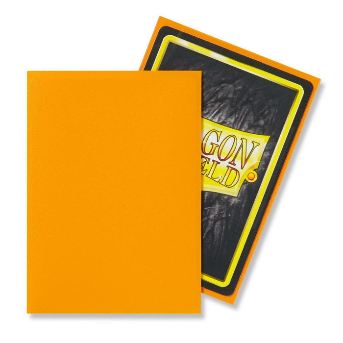 A card with a bright orange front is placed in front of another card. The visible card's back has a black background with the word "DRAGON" in large yellow and red letters, and "SHIELD" in smaller letters at the bottom. Encased in Dragon Shield: Standard 100ct Sleeves - Orange (Matte) by Arcane Tinmen, its edges are outlined in yellow and the border is orange.
