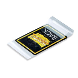 An Arcane Tinmen Dragon Shield: Standard Size 100ct Inner Sleeves - Perfect Fit Sealable (Clear 'Thindra') lies on a white surface. The sleeve is transparent, showing the card inside. The card has a predominantly black color with a yellow and white Dragon Shield logo, and the word "BACK" is visible at the top, indicating that the card is facing down.